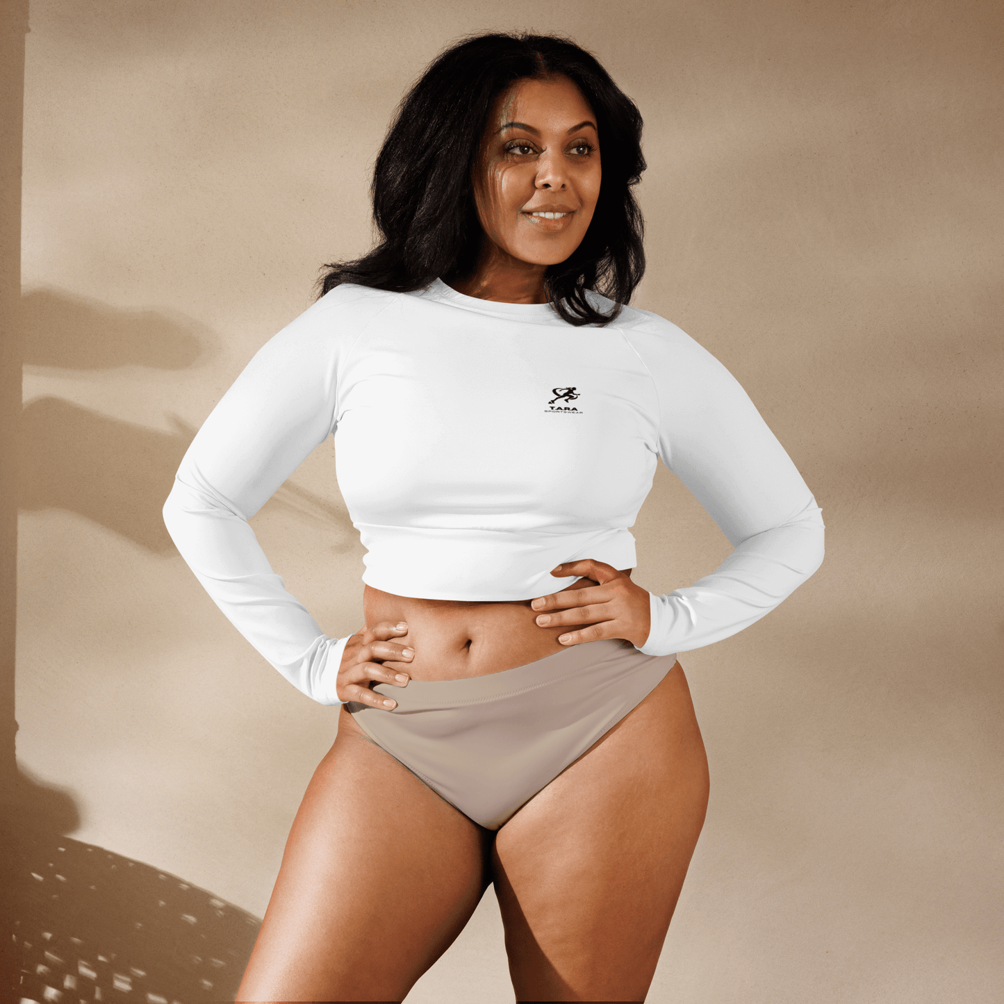 Woman wearing a white long-sleeve crop top and beige high-waisted bikini bottoms against a beige background.