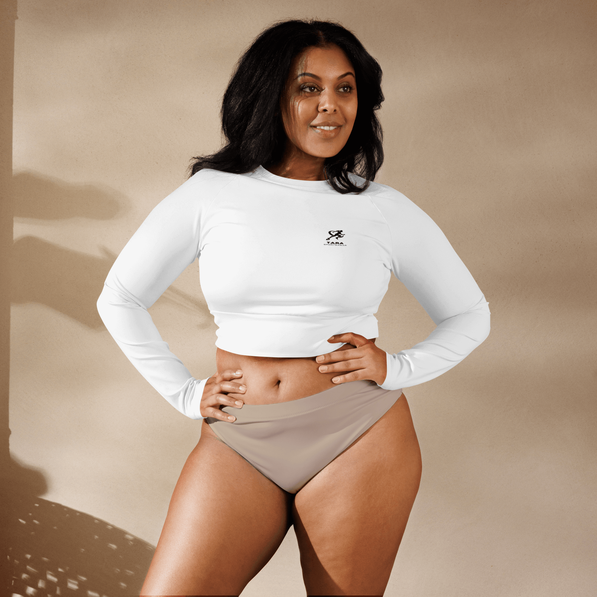 Woman wearing a white long-sleeve crop top and beige high-waisted bikini bottoms against a beige background.