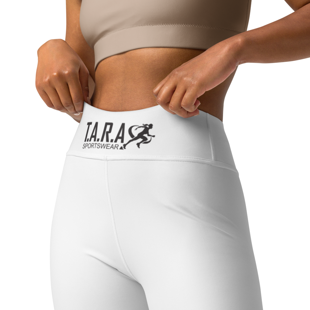 Woman wearing a sports bra and white leggings with a brand logo, standing against a neutral wall.