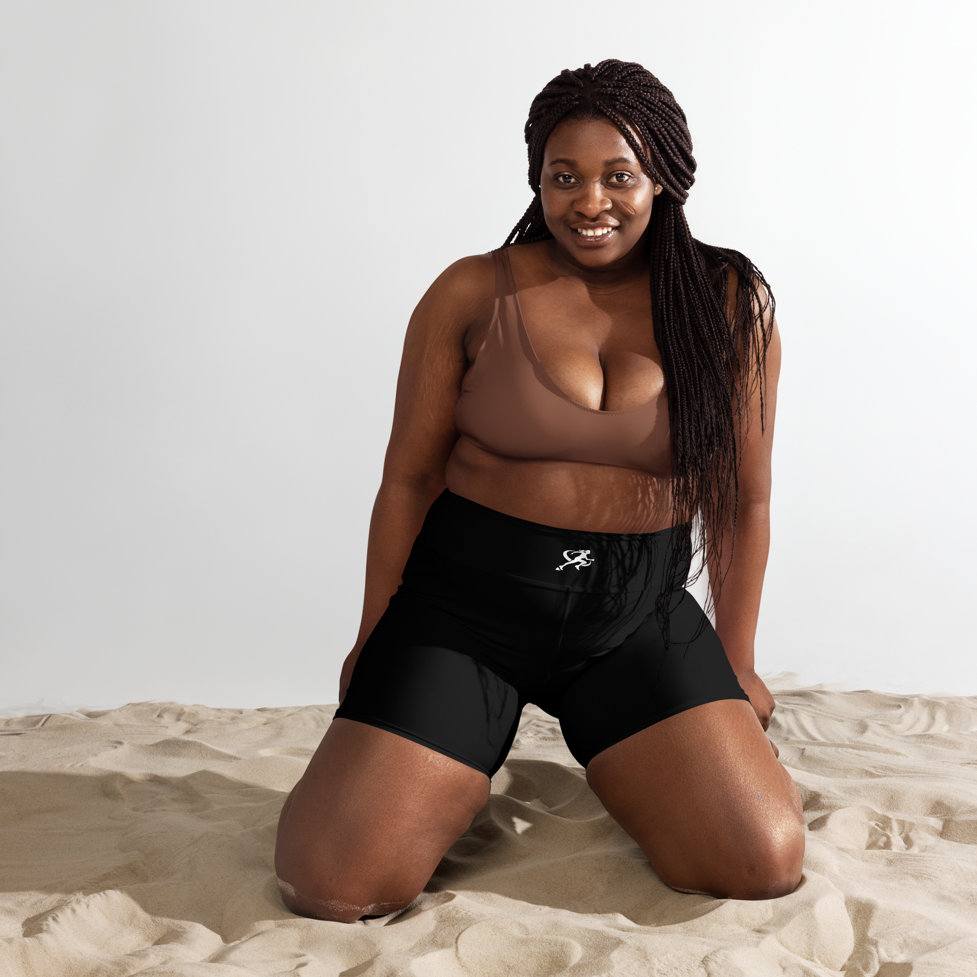 Woman in a yellow top and black shorts standing on sand with an orange sky